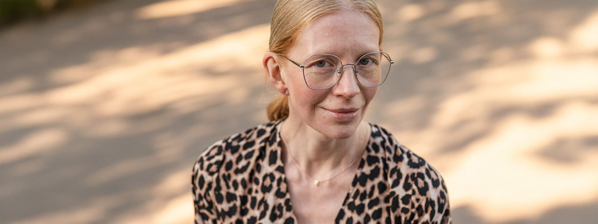 Nainen hymyilee ulkona, kasvoillaan vaaleat silm&auml;lasit